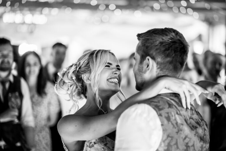 A black-and-white photo of a couple dancing closely at an event at Bartle Hall. The woman is smiling broadly, looking at the man. They are surrounded by a crowd of people, slightly out of focus, with festive lights in the background, capturing a touch of Lancashire charm. Image by Andy Wade Photography.