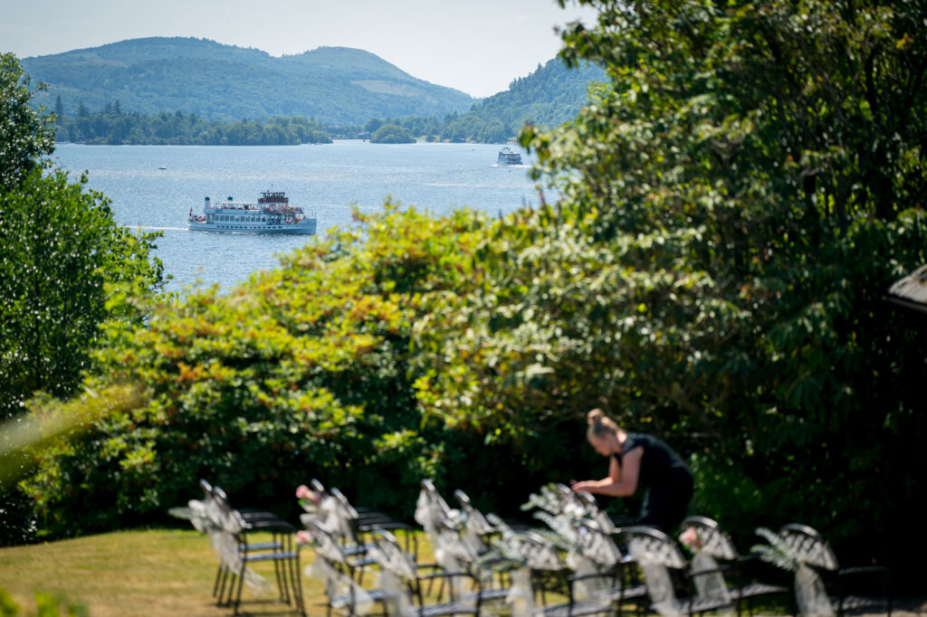 A scenic view of a lake with boats in the distance, surrounded by lush greenery and hills. In the foreground, a person is adjusting decor on chairs arranged for what appears to be an outdoor event. Image by Andy Wade Photography.