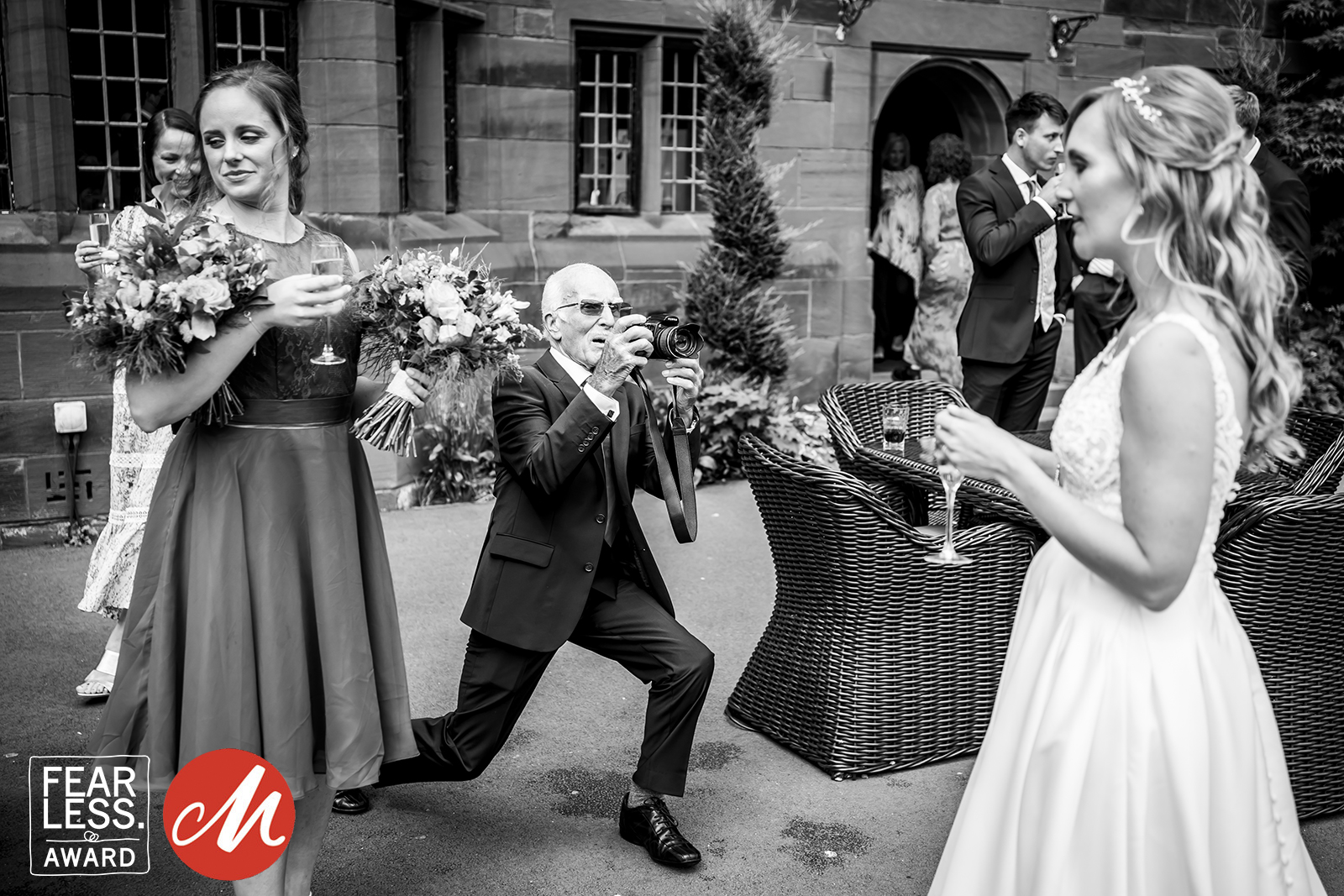 A man in a suit kneels on one knee, holding a glass, while a woman in a bridal dress laughs. Another woman in a dark dress stands nearby holding a bouquet and smiling. The background features a brick building with other guests. "Fearless Award" logo is visible. Image by Andy Wade Photography.