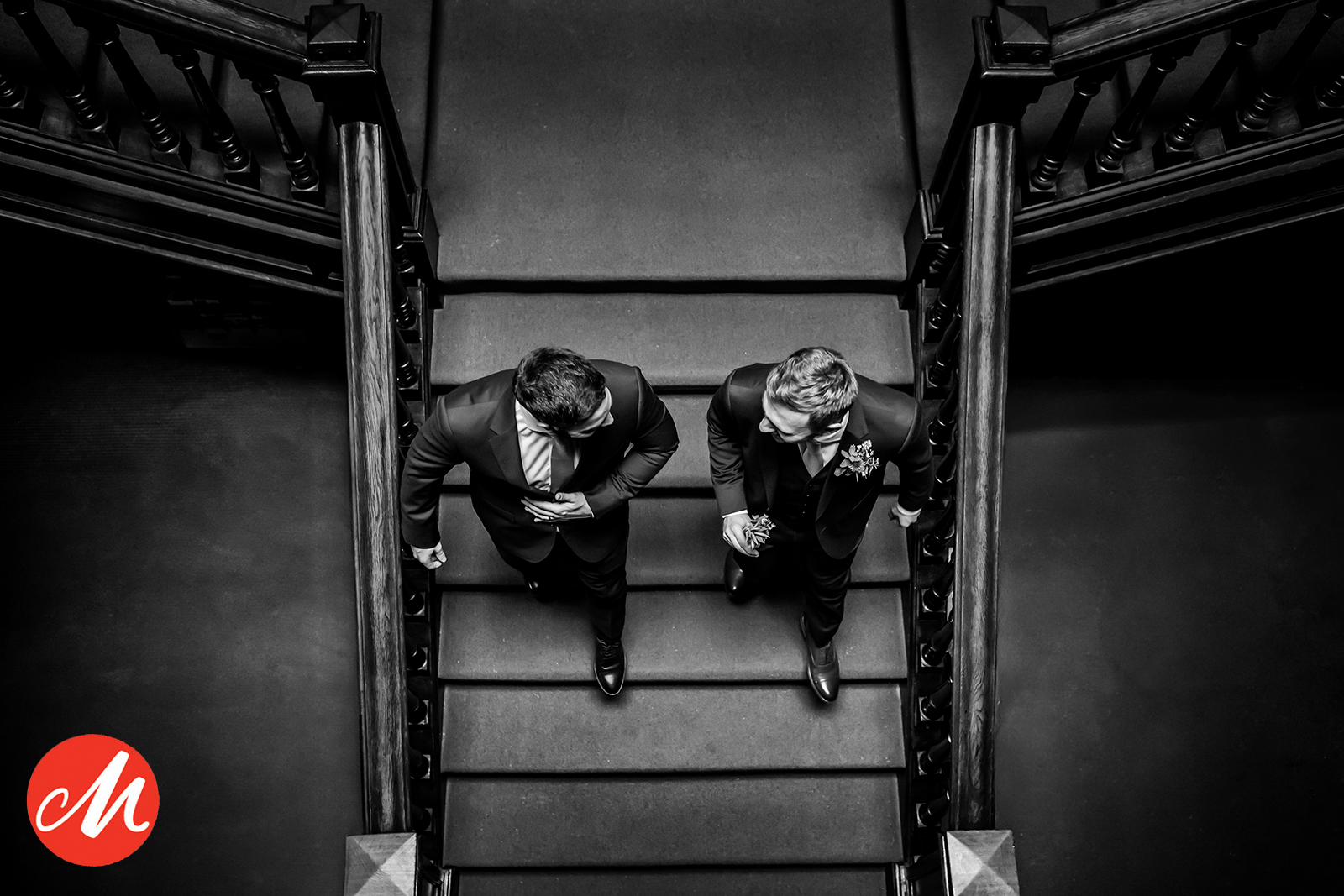 Black and white photo of two men in suits descending a staircase, viewed from above. The man on the right holds a glass, and the man on the left adjusts his tie. A red circular logo with a white "M" is at the bottom left corner. Image by Andy Wade Photography.