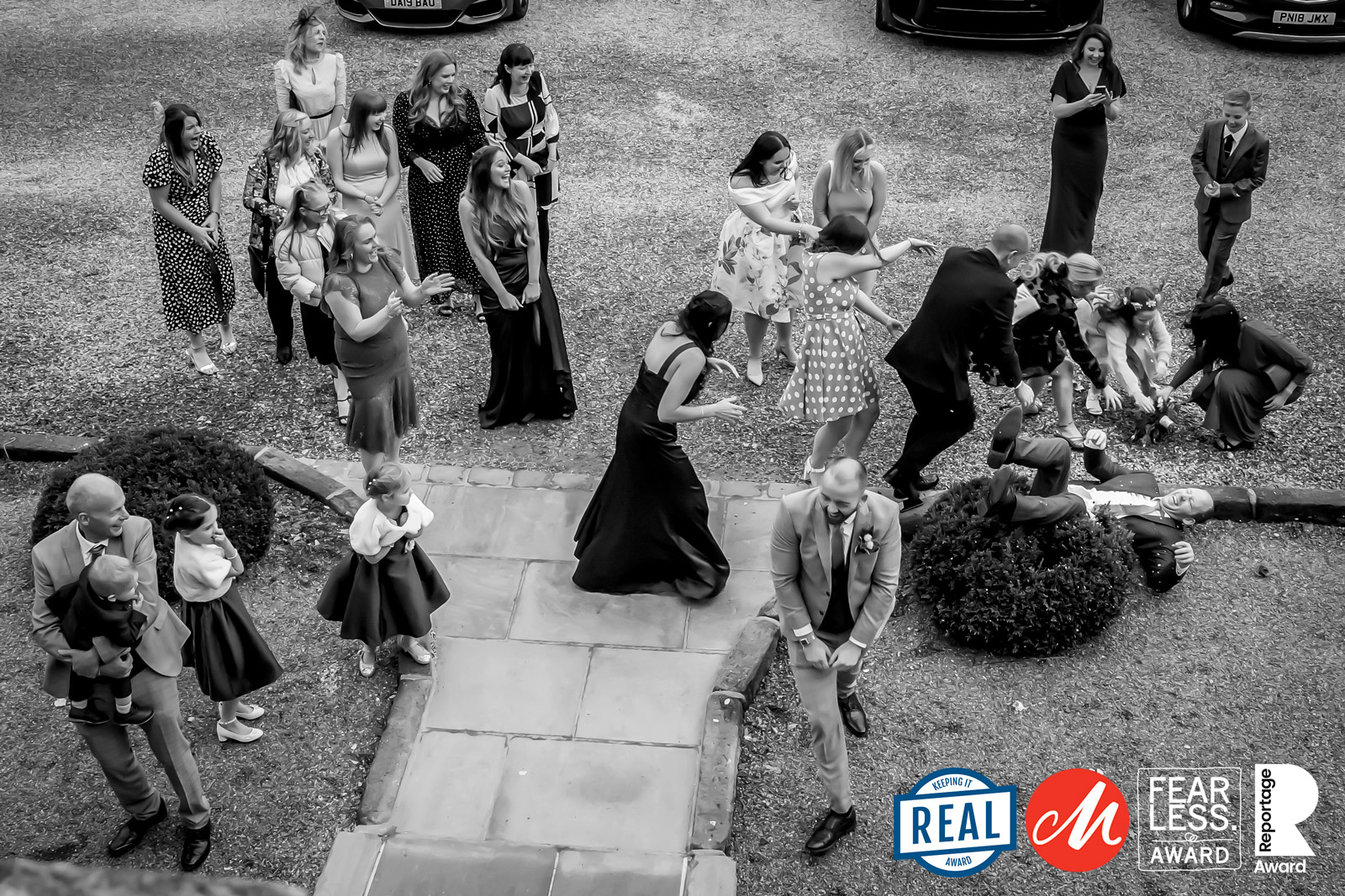 Black and white photo of people gathering outdoors in formal wear. A man lies on the ground playfully while others around him, including a bride, laugh and mingle. Two logos in the bottom right corner: "REAL" and "FEARLESS AWARD - M. Image by Andy Wade Photography.