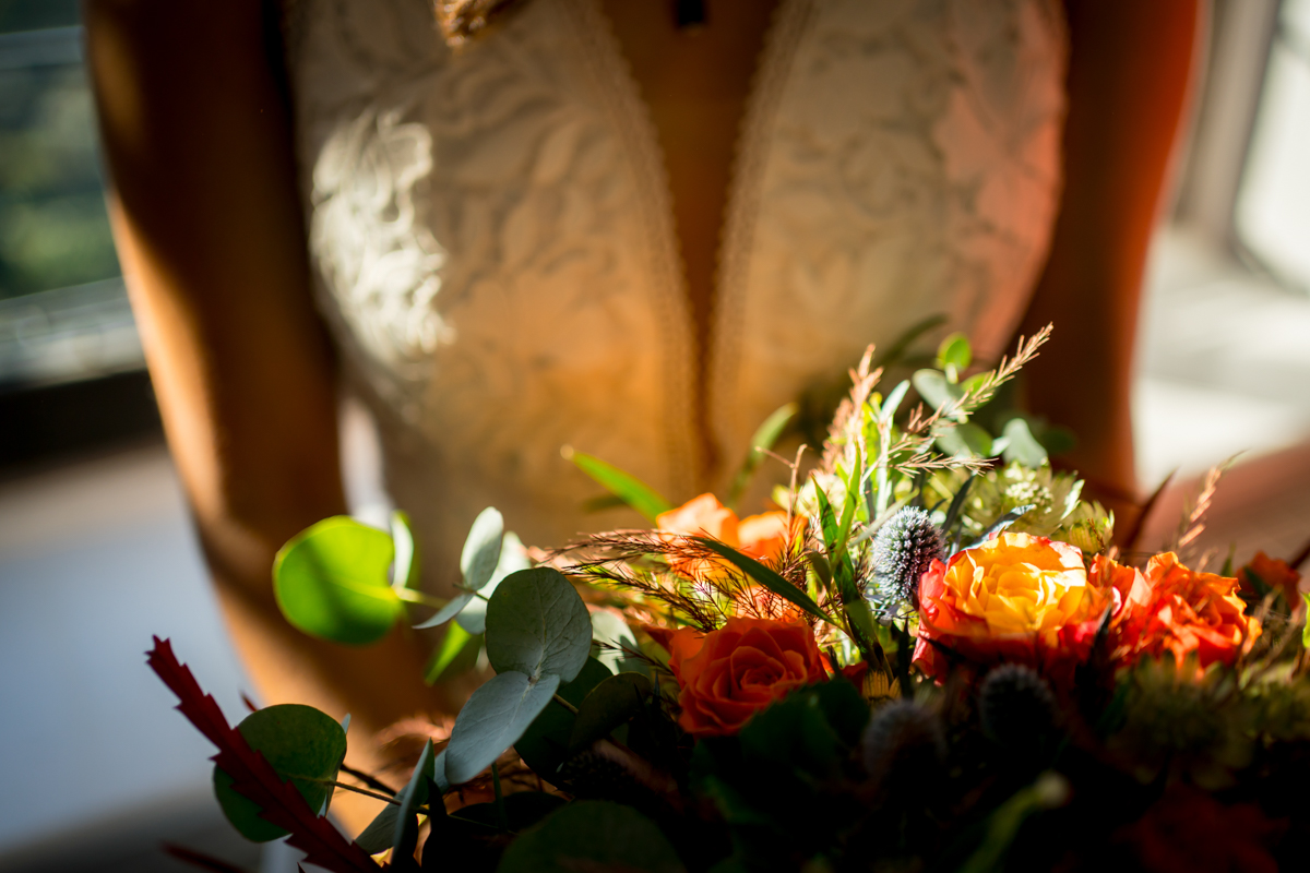 A bride in a lace wedding dress holds a vibrant bouquet of flowers, featuring orange roses and green foliage. Sunlight streams in, highlighting the details of the dress and flowers, creating a warm and intimate atmosphere. Image by Andy Wade Photography.