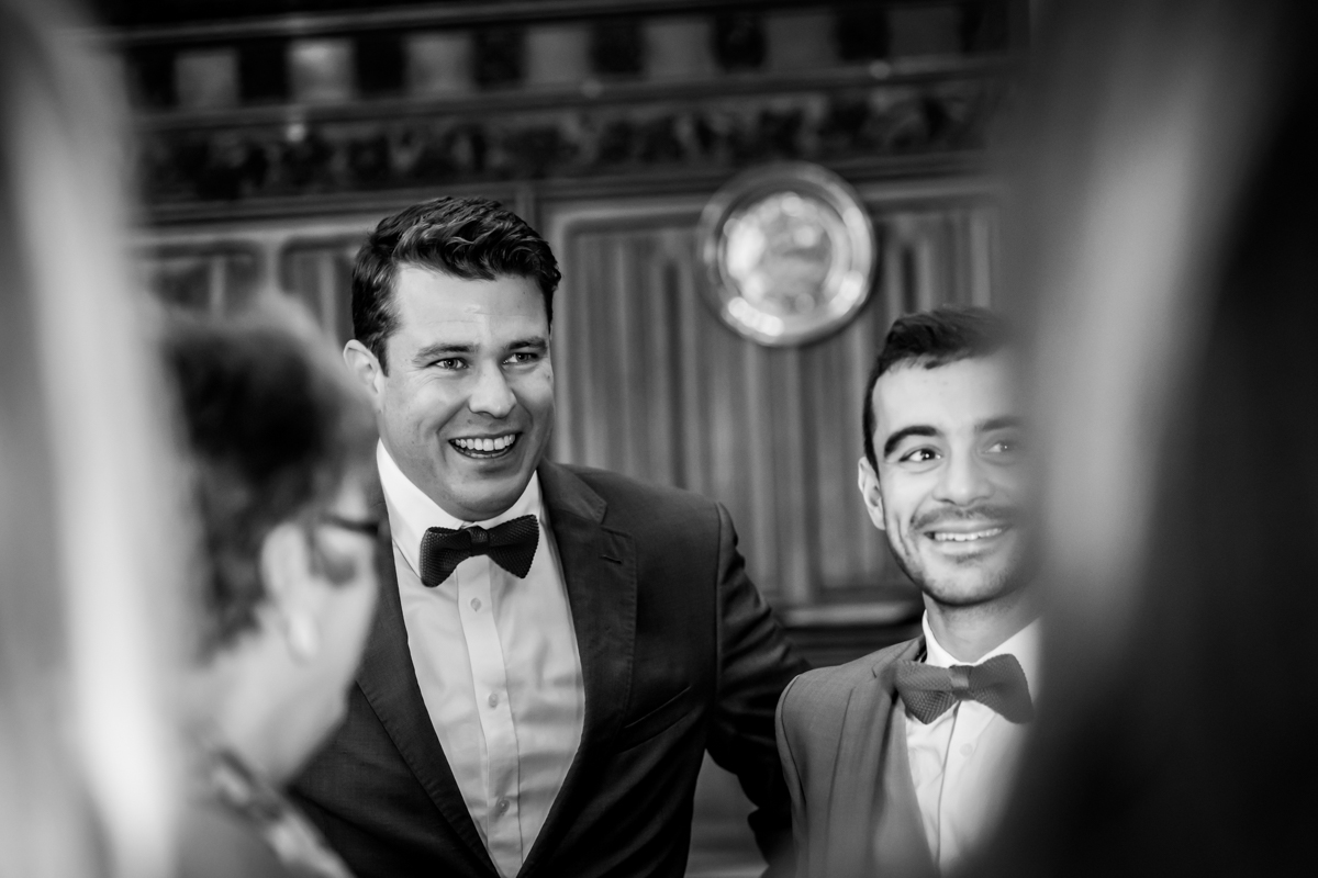 A black-and-white photo showing two men in formal attire; one on the left with a big smile wearing a bow tie and a dark suit, and the other man to the right, also smiling, in a lighter suit and bow tie. They are surrounded by people, with a wooden wall and a circular emblem in the background. Image by Andy Wade Photography.