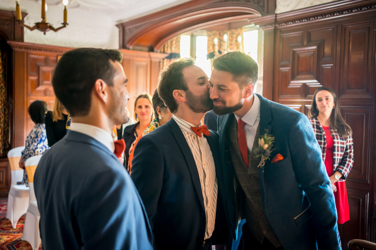 Two men, both in suits with one in a blue blazer and the other in a dark blazer with a bow tie, share a kiss in a warmly lit room. Several people in colorful attire stand in the background, watching with smiles. The room features dark wood paneling and ornate decor. Image by Andy Wade Photography.