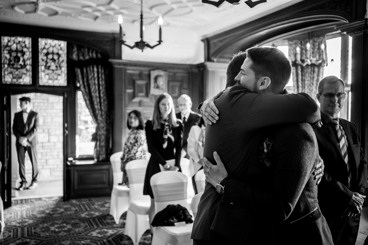 A black and white photo of two men embracing in a warmly lit room during a ceremony. They are surrounded by seated attendees who are smiling and watching them. The background includes large windows with stained glass and ornate wooden decor. Image by Andy Wade Photography.