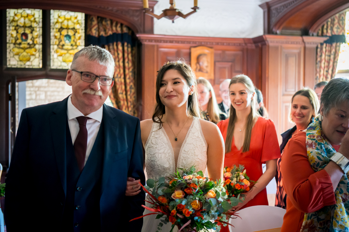 A smiling bride in a white dress holds a vibrant bouquet of flowers as she walks down the aisle with an older man in a dark suit and tie. Behind them, joyful guests in colorful attire follow, with wood-paneled walls and stained-glass windows in the background. Image by Andy Wade Photography.