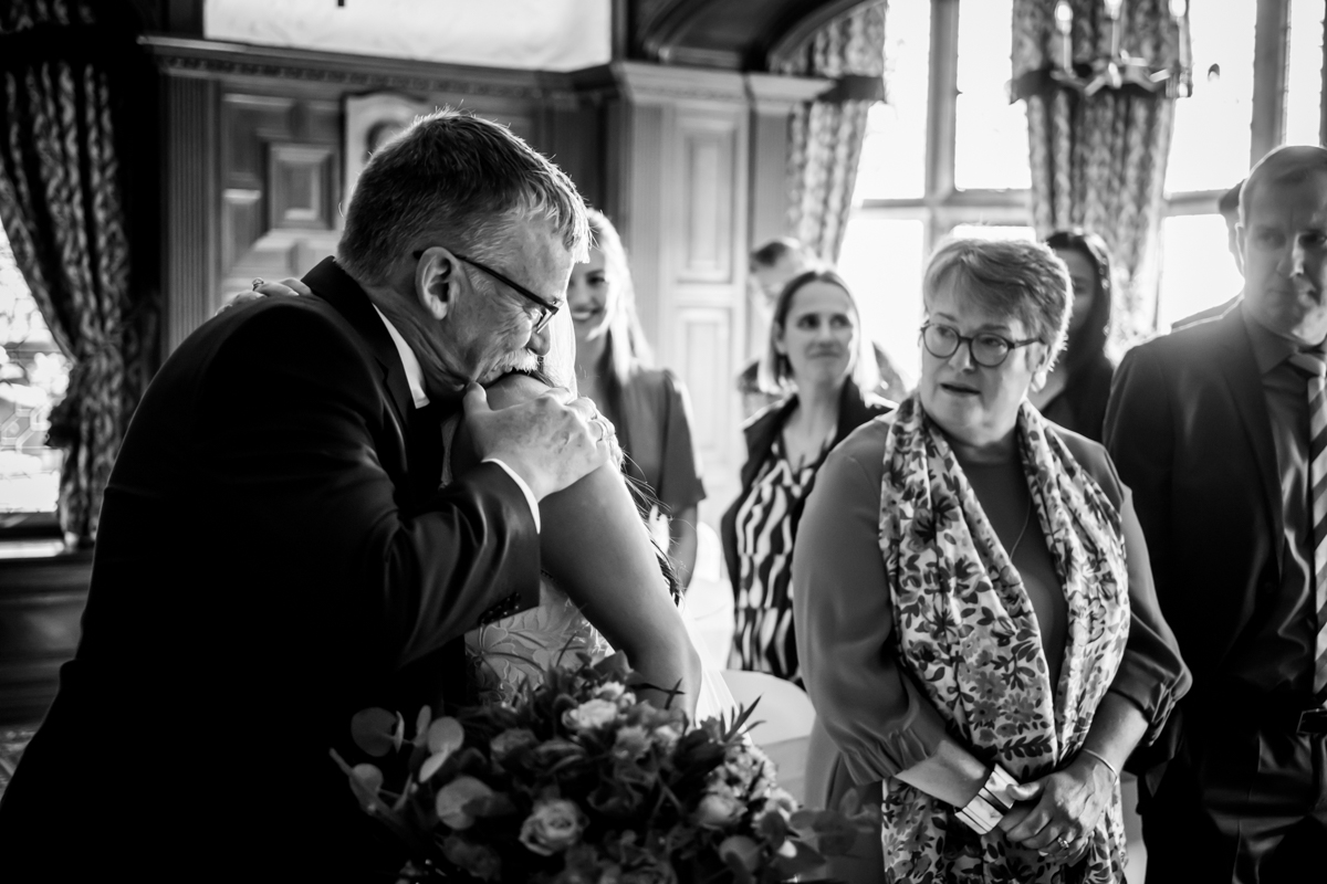 A bride embraces an older man in a warm hug inside an elegant room. The bride holds a bouquet. Several people stand nearby, watching the interaction, including a woman with glasses and a patterned scarf, who appears touched by the moment. Image by Andy Wade Photography.