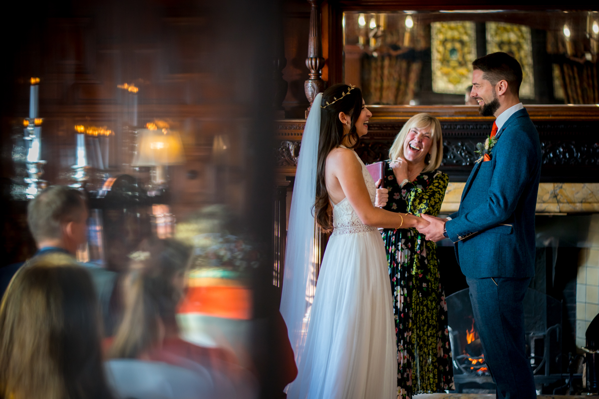 A bride in a white dress and a groom in a blue suit hold hands and smile at each other during their wedding ceremony, officiated by a woman in a floral dress. Attendees are seated, and the venue has warm, elegant decor with candles and a fireplace in the background. Image by Andy Wade Photography.