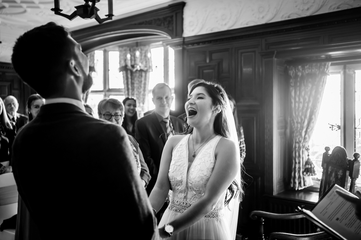 A bride and groom share a joyful moment during their wedding ceremony in a vintage-style room. The bride is laughing heartily while holding the groom's hands. Guests in the background are smiling, and natural light pours in through large windows. Black and white photo. Image by Andy Wade Photography.