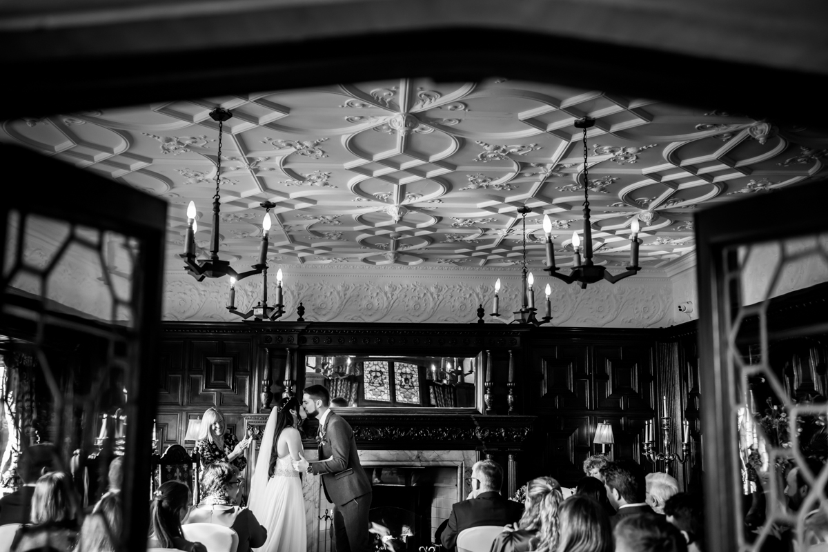 A couple stands at the altar under an ornate ceiling, holding hands and gazing at each other during their wedding ceremony. Guests seated in chairs witness the moment in a richly decorated room with chandeliers and a large mirror above the fireplace. Image by Andy Wade Photography.
