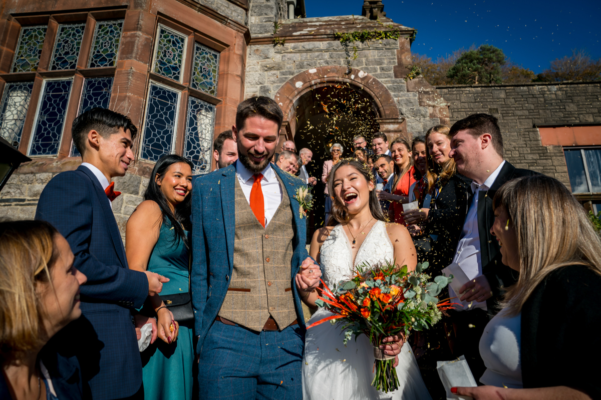 A newlywed couple is joyfully walking down steps outside a stone building, surrounded by cheering guests throwing confetti. The bride, in a white dress, is holding a bouquet of orange flowers, and the groom is wearing a blue suit with an orange tie. The sky is clear and blue. Image by Andy Wade Photography.