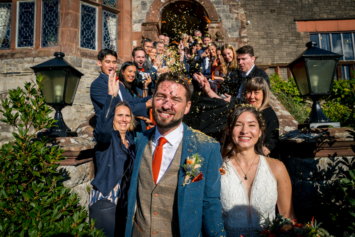 A bride and groom smile brightly as they walk down steps while guests throw confetti at them. The bride wears a white dress and the groom is in a blue suit with an orange tie. The background features a stone building with a dark wooden door and stained glass windows. Image by Andy Wade Photography.