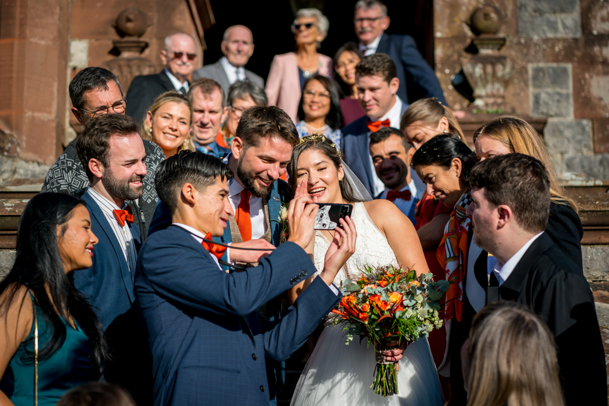 A group of people gathered on the steps of a building, posing for a photo at a wedding. The bride, holding a bouquet of flowers, and the groom, both in front, are smiling and looking at a phone held by another guest. The group appears joyful and celebratory. Image by Andy Wade Photography.