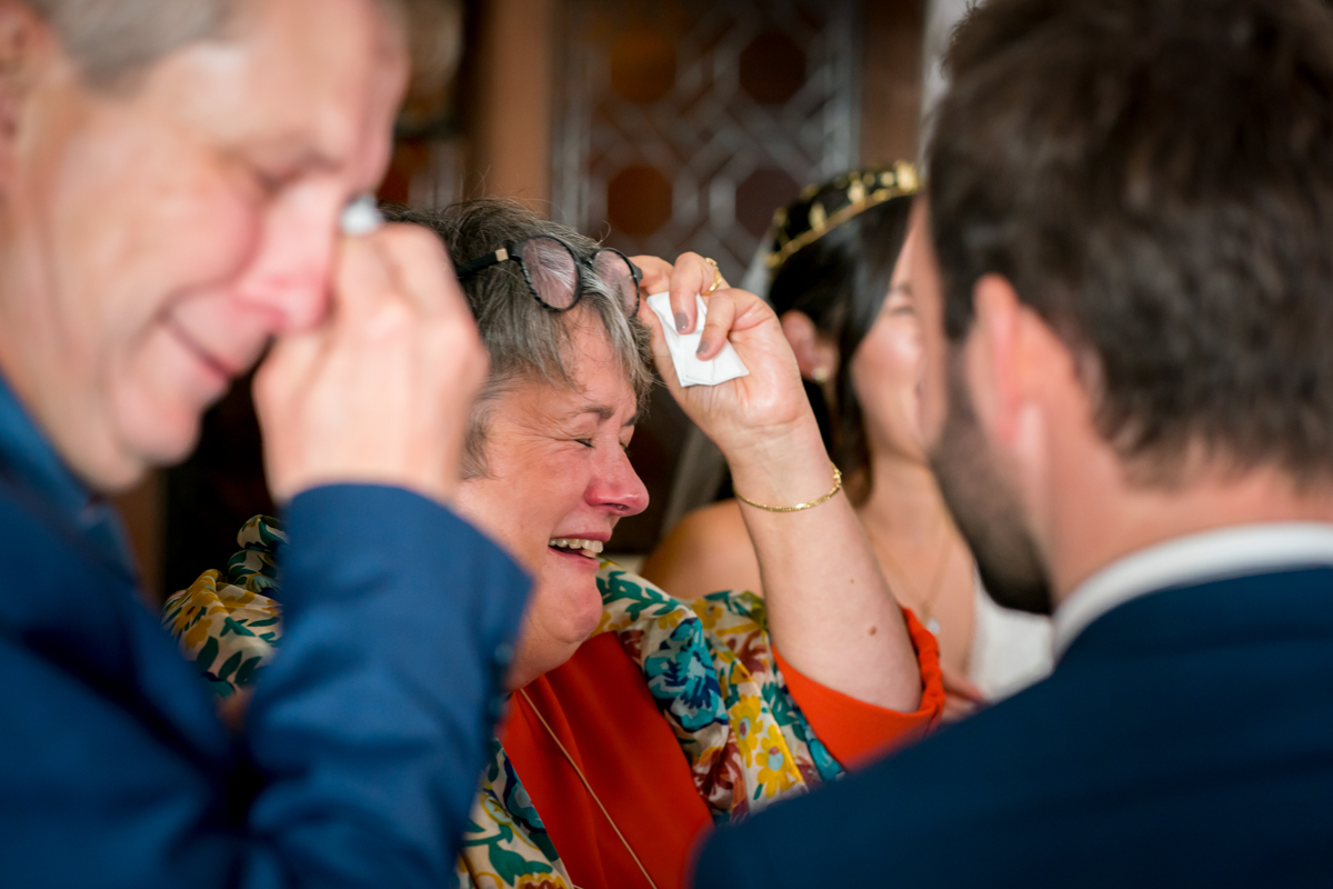 An older woman in colorful clothes wipes away tears and laughs with two men and a woman in the background. The group appears to be sharing a joyful moment indoors, possibly at a celebratory event. Image by Andy Wade Photography.