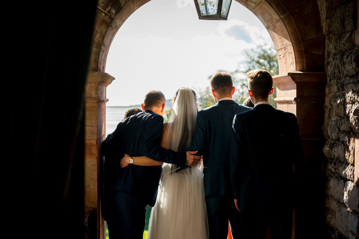 A bride in a white dress and veil stands with four men in suits under a stone archway. They face away from the camera, looking outside into the bright daylight. The men have their arms around each other, creating a feeling of closeness and camaraderie. Image by Andy Wade Photography.