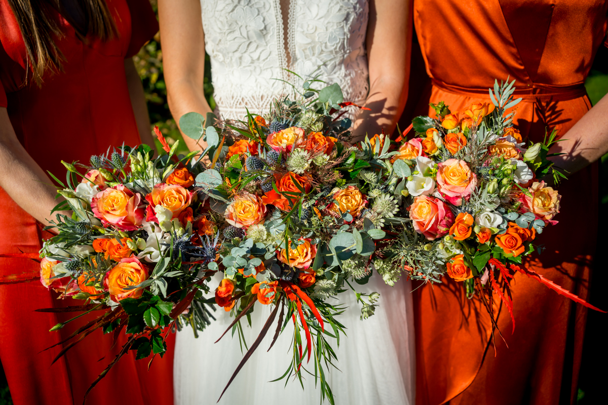 Three women in orange and white dresses holding vibrant floral bouquets composed of orange roses, green foliage, and assorted colorful flowers. The central figure is in a white gown, flanked by women in orange. The scene is outdoor with bright lighting. Image by Andy Wade Photography.