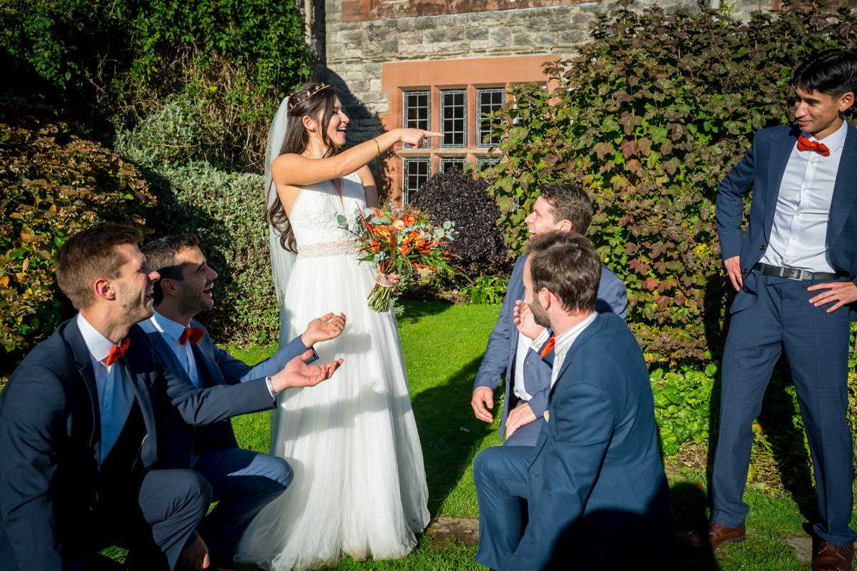 A bride in a white dress and holding a bouquet is standing and pointing while smiling at four groomsmen kneeling or crouching around her. They are all dressed in blue suits with red bow ties and are in front of a building with stone and red brick walls. Image by Andy Wade Photography.