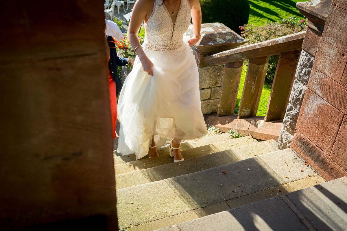 A bride in a white wedding dress ascends stone steps outside, holding her dress up to avoid tripping. Another person in formal attire walks closely behind her, partially visible. Sunlight casts shadows on the stairs and surrounding green garden. Image by Andy Wade Photography.