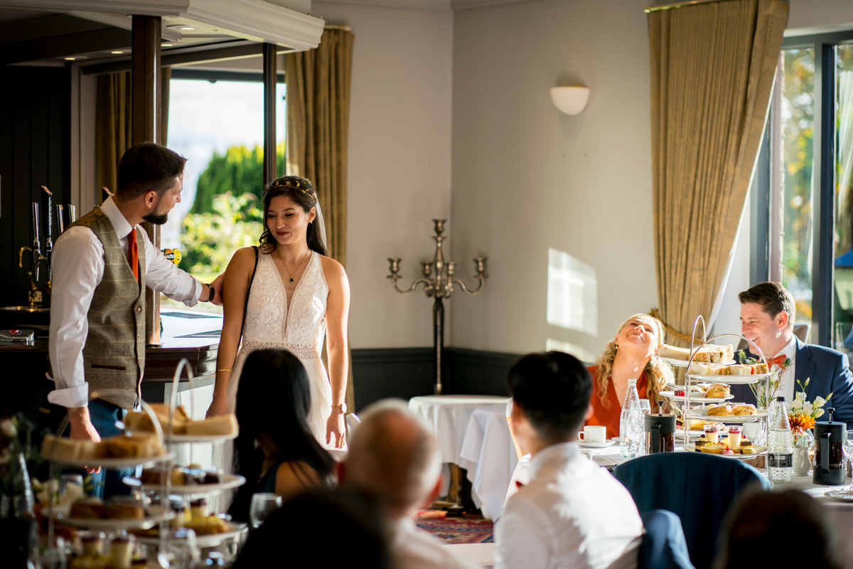 A groom, wearing a vest and tie, stands and gestures towards his bride, who is in a white dress and smiling. Guests are seated around tables set with tiered trays of food, attentively watching and smiling. The room is warmly lit with natural light from large windows. Image by Andy Wade Photography.