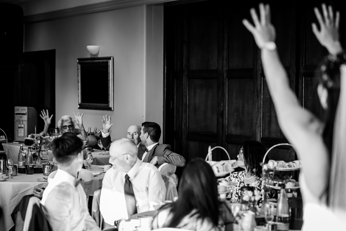A black and white photo of a lively reception or dinner gathering. People are seated at round tables, interacting and raising their hands excitedly. One person in the foreground has both arms raised. The room has wooden paneling and a framed mirror on the wall. Image by Andy Wade Photography.
