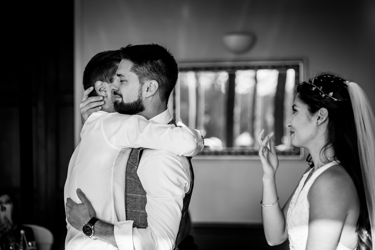 A black and white photo shows two men embracing warmly while a woman in a wedding dress watches with a smile. The room has a softly blurred background with a large mirror reflecting light. Image by Andy Wade Photography.