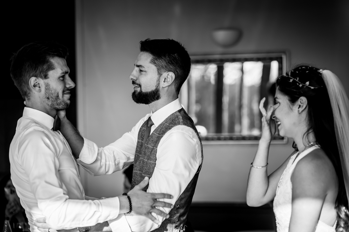 A groom holds and looks at the best man with emotion, both dressed in formal attire. A bride in a white gown and veil stands to the side, smiling and dabbing her eyes with her hand. The scene appears heartfelt, likely taking place during a wedding ceremony. Image by Andy Wade Photography.