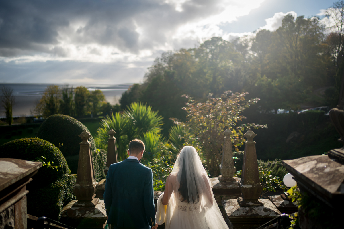 A bride and groom stand on a stone staircase, facing a scenic landscape with lush greenery and a distant body of water under a partly cloudy sky. Backlit by sunlight, the couple appears in a serene, picturesque moment of their wedding day. Image by Andy Wade Photography.
