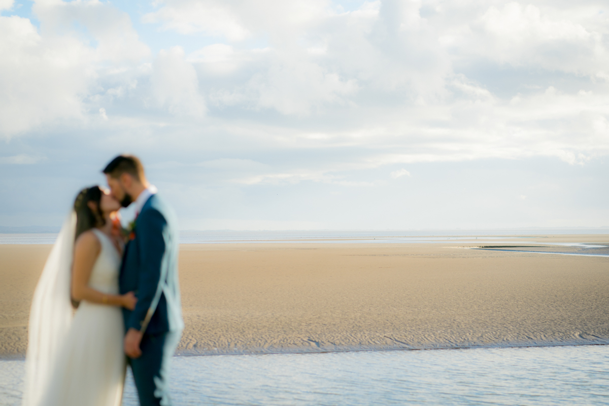 A couple in wedding attire shares a kiss on an empty beach with an expansive view of sand, blue sky, and soft clouds in the background. The bride is wearing a white dress and veil, while the groom is in a blue suit. The image is slightly blurred, creating a dreamy effect. Image by Andy Wade Photography.