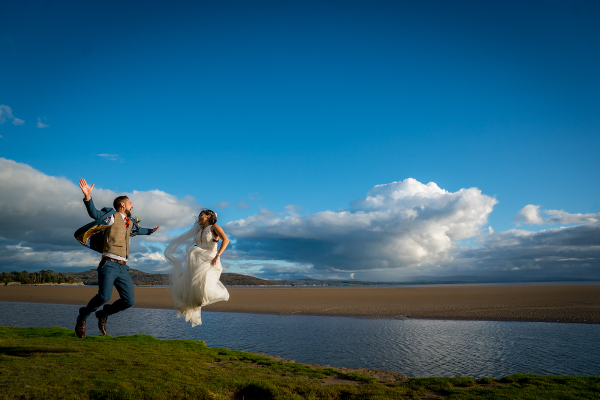 A bride and groom jump joyfully in the air on a grassy area by the beach, with a bright blue sky and fluffy clouds in the background. The bride is in a white dress, and the groom is in a suit with a vest. The ocean and sandy beach stretch out behind them. Image by Andy Wade Photography.