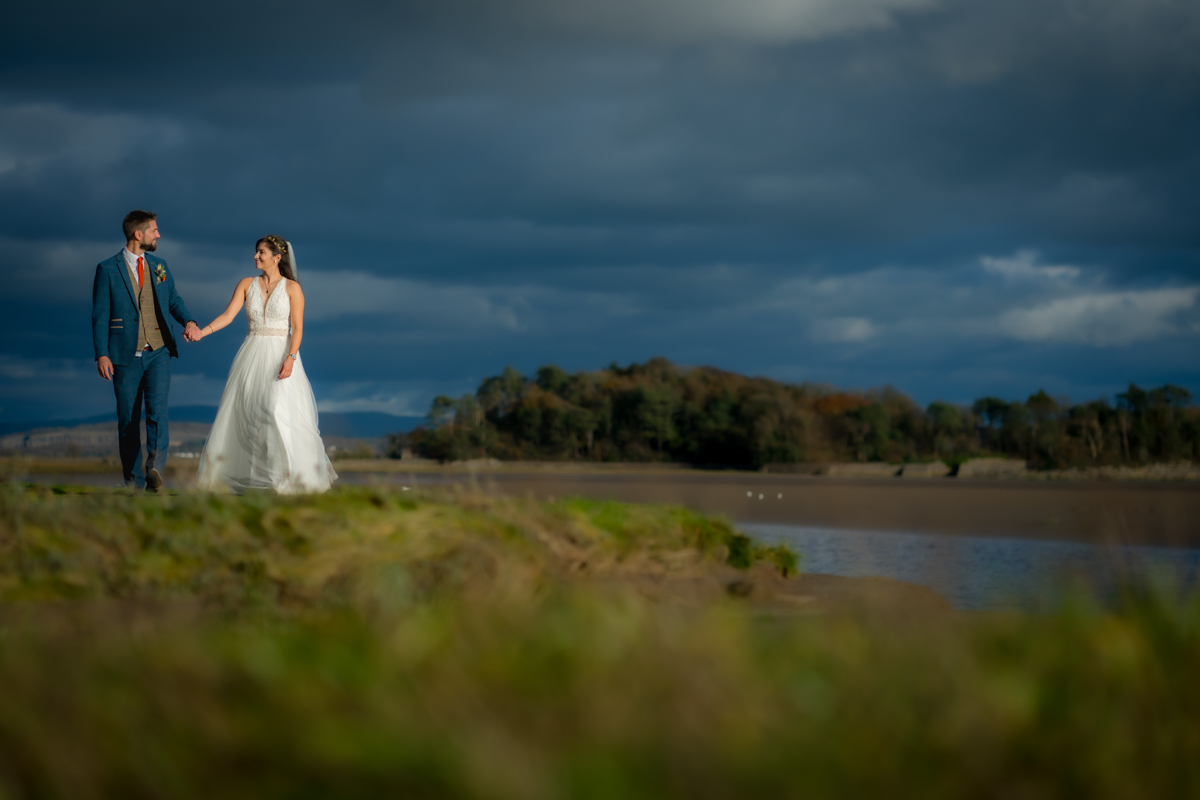 A bride and groom hold hands while walking outdoors. They are dressed in wedding attire; the bride in a white gown and the groom in a suit. They are on a grassy path near a body of water with a scenic landscape and cloudy sky in the background. Image by Andy Wade Photography.