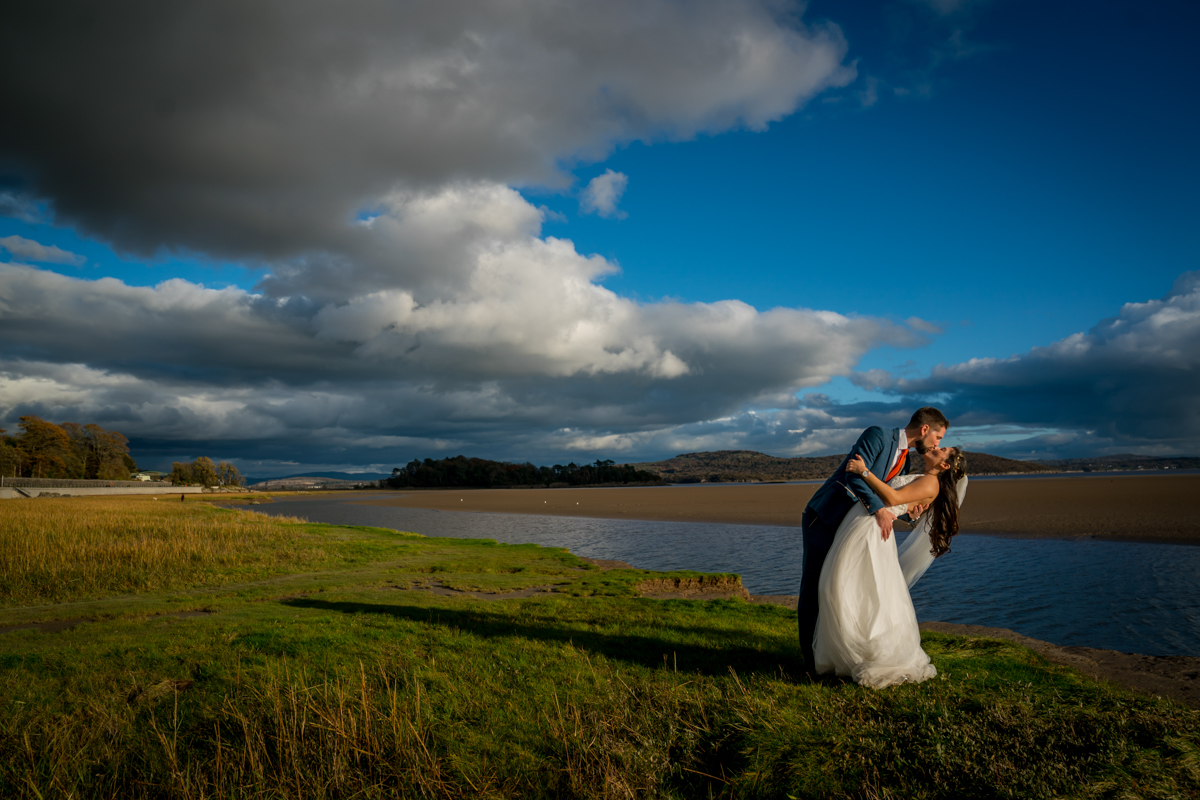 A bride and groom share a kiss while the groom dips the bride on a grassy bank by a calm lake under a dramatic cloudy sky. The bride wears a flowing white dress, and the groom is dressed in a blue shirt and trousers. Image by Andy Wade Photography.