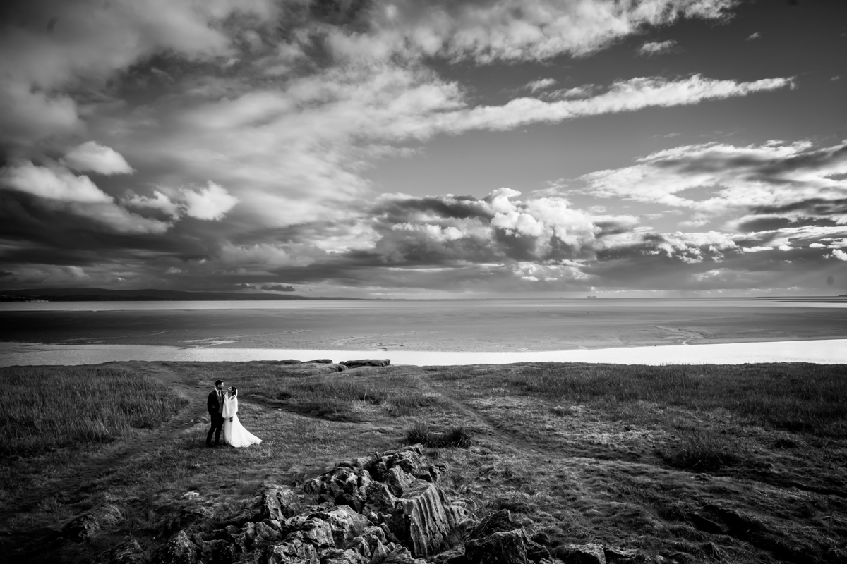 A black-and-white photo of a bride and groom standing together on a grassy cliffside. They face away from the camera, looking out over a vast landscape with dramatic clouds and distant water. Rocky terrain and patches of grass are visible in the foreground. Image by Andy Wade Photography.