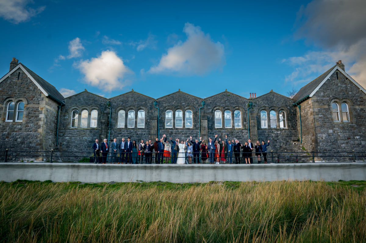 A large group of people dressed formally stand in front of a historic stone building with arched windows, posing for a photo on a sunny day. The foreground features tall grass, and the blue sky with a few clouds creates a picturesque backdrop. Image by Andy Wade Photography.