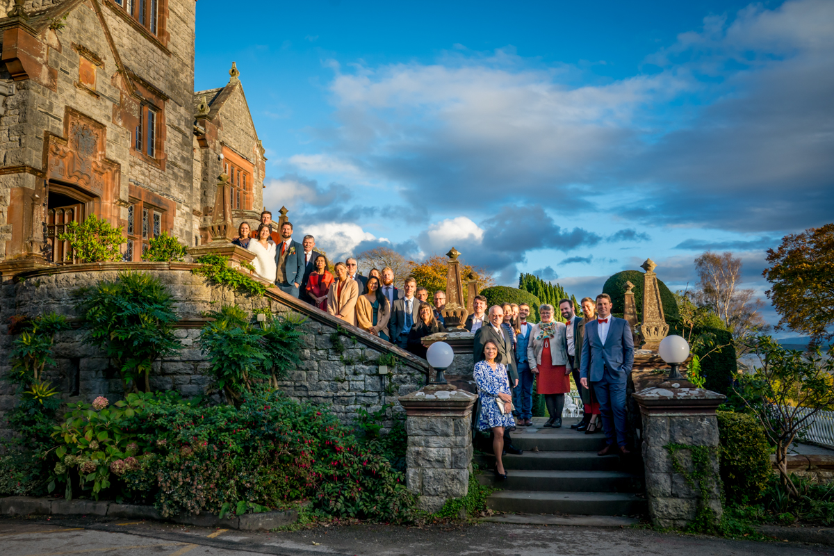 A group of people dressed in formal attire poses on the stone steps and terrace of a large, historic building. The setting includes lush gardens and distant trees with a blue sky and scattered clouds. It appears to be late afternoon or early evening. Image by Andy Wade Photography.