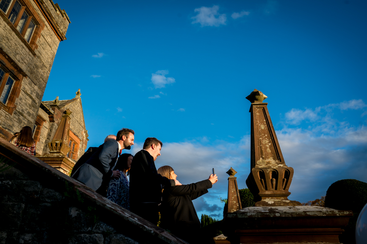 A group of four people stands on the steps of an old building with stone architecture, taking a selfie as the sun sets, casting warm light and shadows. The sky is clear and blue with a few clouds. They appear to be enjoying the moment together. Image by Andy Wade Photography.