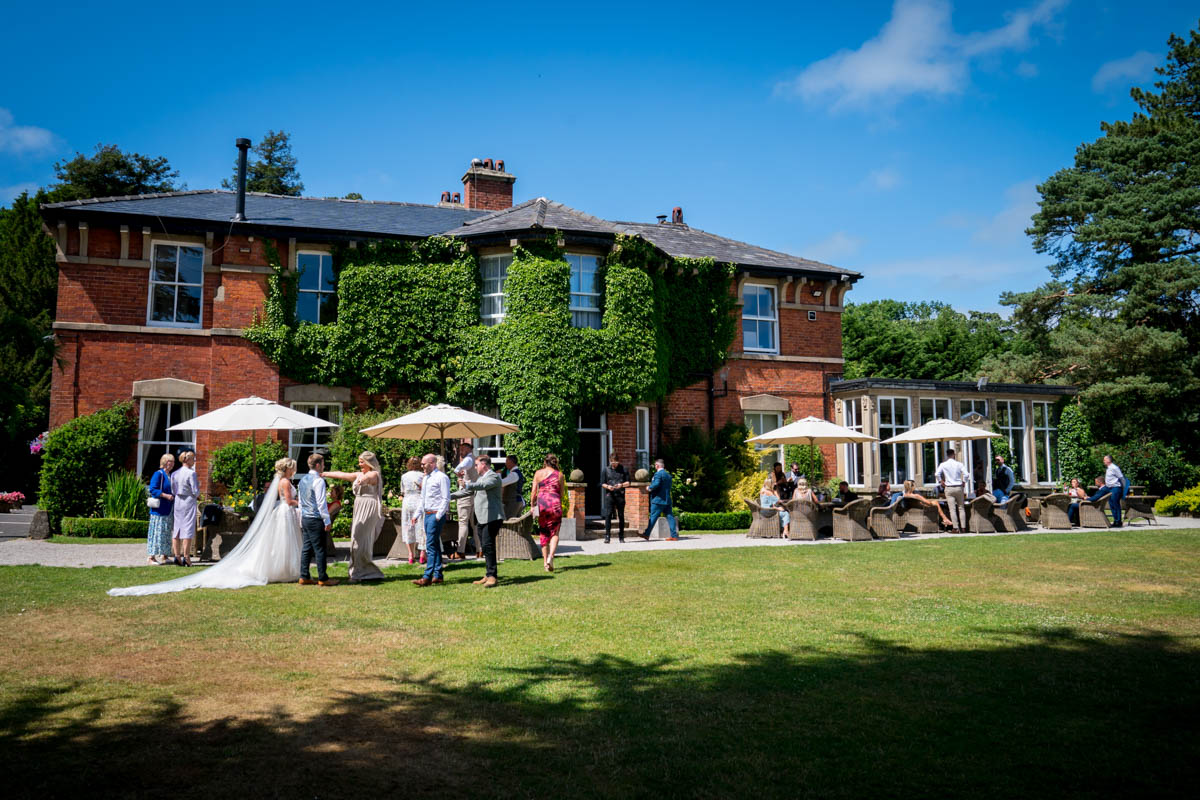 A group of people are gathered outside a large, two-story brick building covered in ivy. The scene appears to be a wedding reception, with guests dressed in formal attire standing on a lawn with parasols and seating areas. A photographer captures charming moments against the backdrop of the clear, blue sky. Image by Andy Wade Photography.