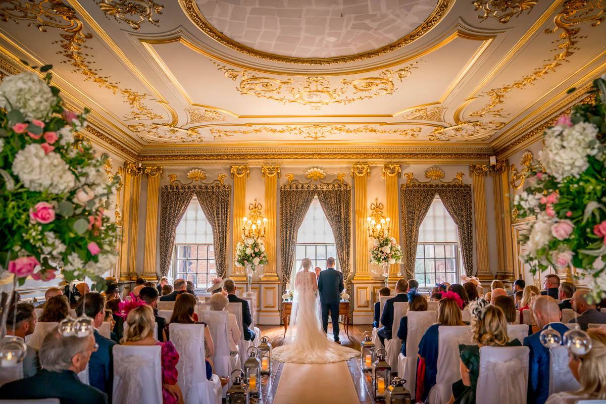 A bride and groom stand at the altar in an ornate, gold-themed room with high ceilings and large windows. Guests are seated in rows facing the couple, while a Lancashire wedding photographer captures every moment. The room is adorned with floral arrangements and elegant drapery, creating a luxurious ambiance. Image by Andy Wade Photography.