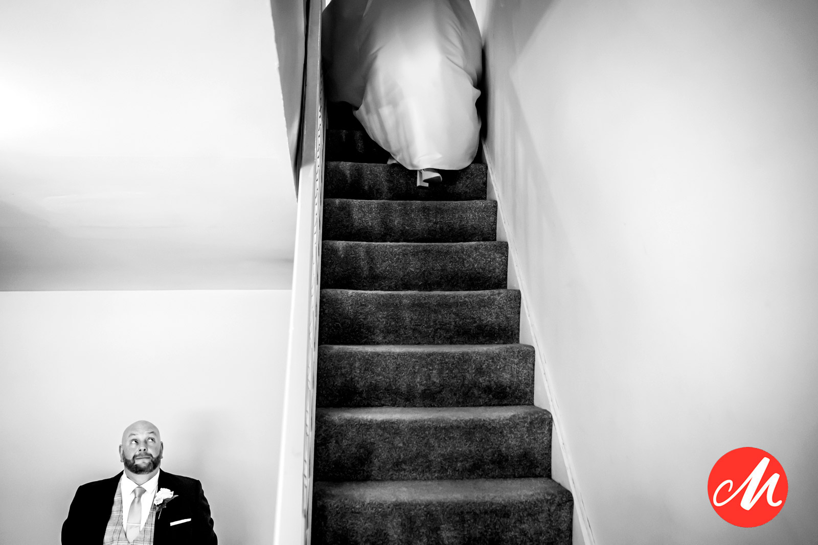 A black and white photo of a person dressed in formal wear sitting at the bottom of a staircase, seemingly looking up. Another person in a gown is captured from behind climbing the stairs. A red circular logo with the letter "M" is in the bottom right corner. Image by Andy Wade Photography.