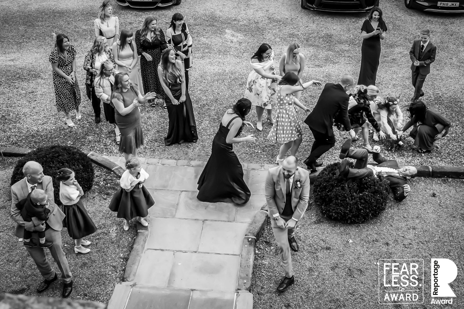 A group of people in formal attire is gathered outside on a stone walkway. Some are seated on the ground, while others stand or walk around. The image portrays a candid moment with people in various playful and engaged poses. A "Fearless Award" logo is visible. Image by Andy Wade Photography.