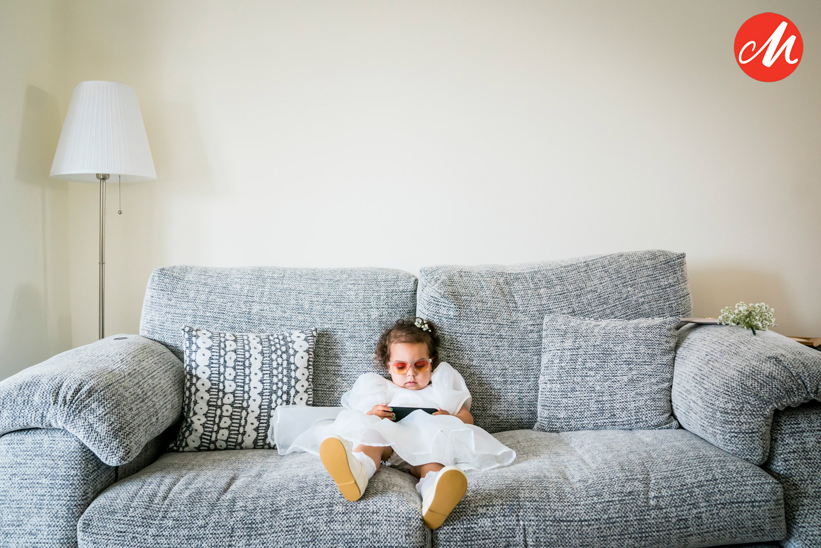 A young girl wearing a white dress and pink sunglasses sits on a grey sofa, holding a black electronic device. The sofa has two patterned cushions, and there is a floor lamp and a small table with plants in the corner of the room. A red "M" logo is in the top right corner. Image by Andy Wade Photography.
