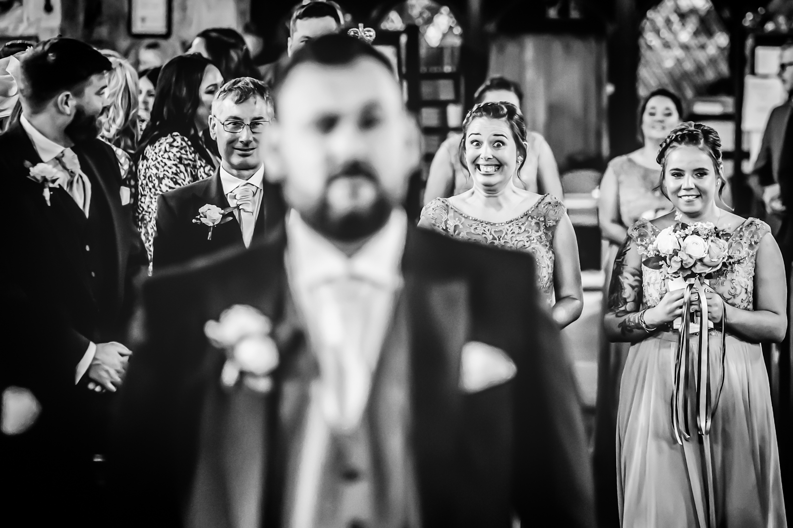 A black and white image captures a wedding ceremony. The groom, slightly out of focus in the foreground, faces a group of guests. A smiling woman in a dress holds a bouquet, standing next to another smiling woman like cherished assets in this emotional portfolio. Other guests are seated and standing in the background. Image by Andy Wade Photography.