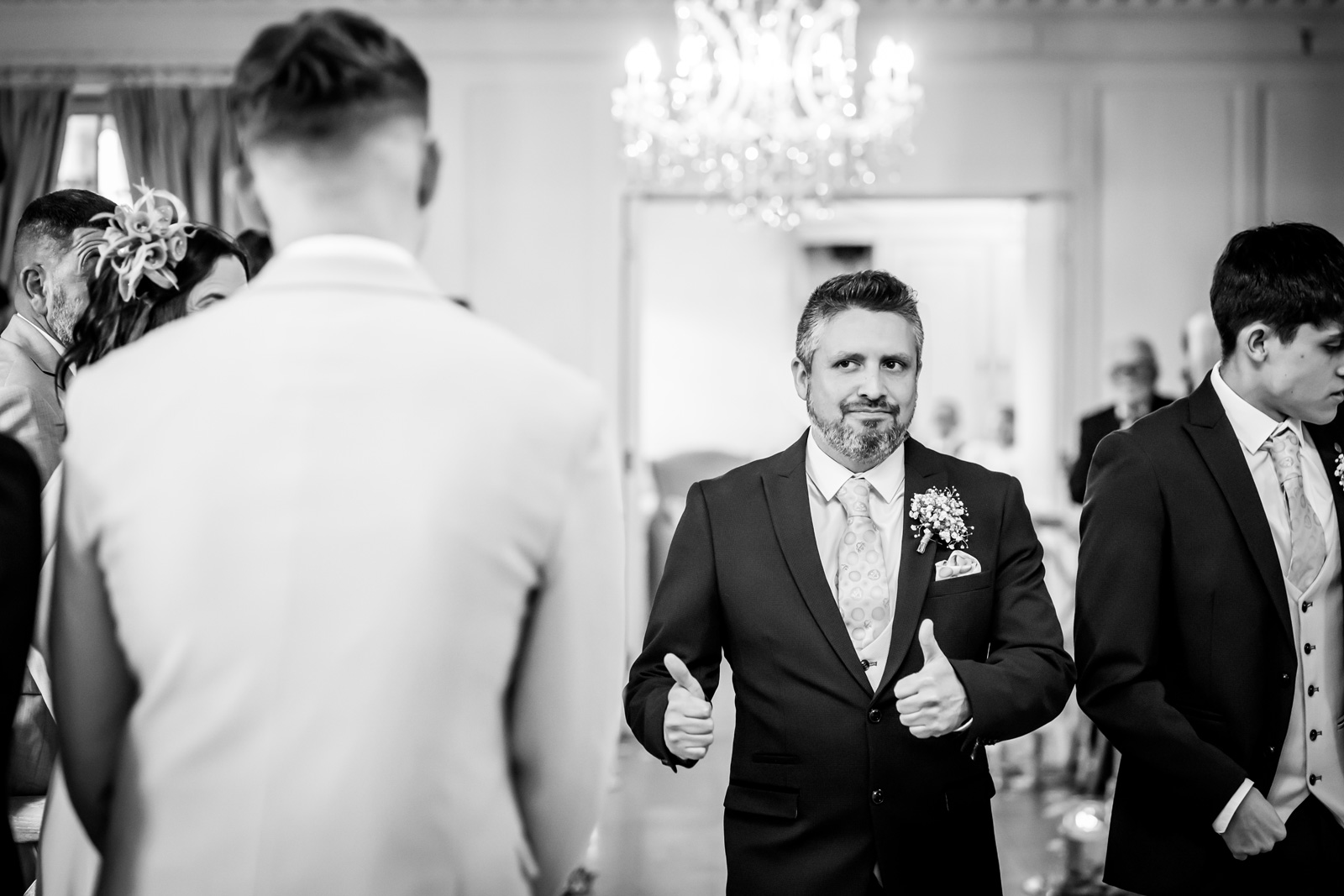 A man in a suit gives two thumbs up while standing in a formal setting with chandeliers and elegant decor, perfect for an impressive portfolio shot. He has a boutonniere on his lapel. Other formally dressed individuals are seen partially in the background and foreground. The scene looks celebratory. Image by Andy Wade Photography.