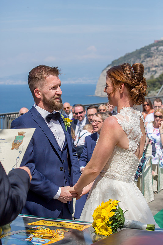 A bride and groom stand facing each other and holding hands during their wedding ceremony by a scenic seaside location. The groom is in a navy suit, and the bride is in a lace wedding dress. Guests are seated behind them, and there is a document in the foreground. Image by Andy Wade Photography.
