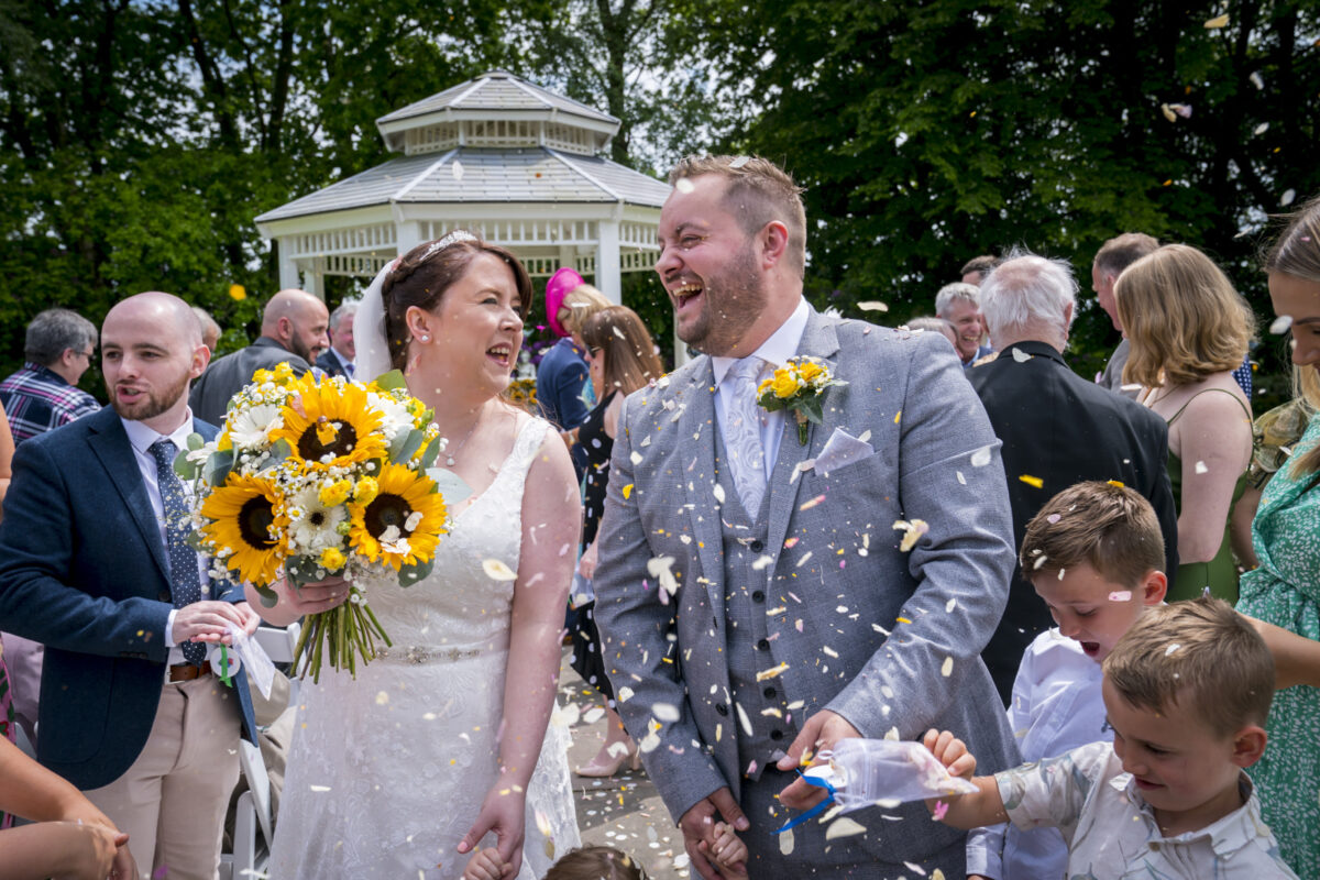 A bride and groom smile joyfully while being showered with confetti at their outdoor wedding at The Higher Trapp Hotel in Lancashire. The bride holds a bouquet of sunflowers and white flowers, and guests surrounding them share in the celebration. A white gazebo is in the background. Image by Andy Wade Photography.