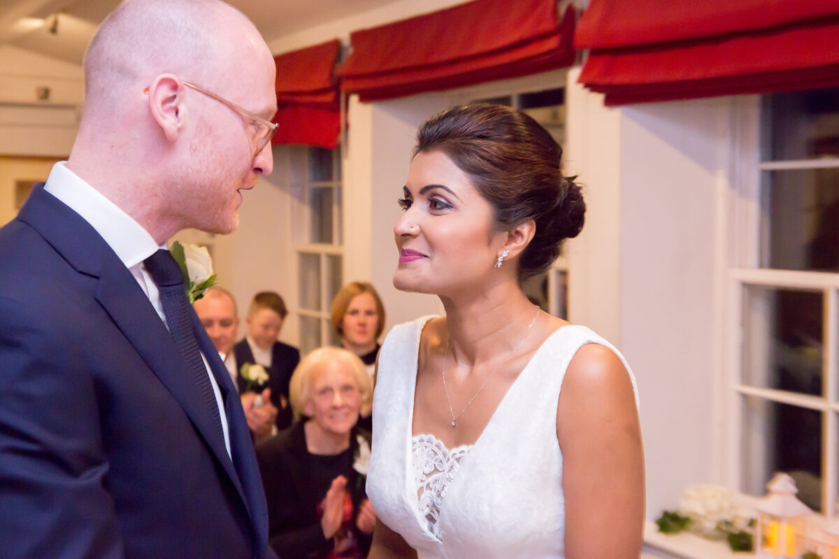 A couple stands together at what appears to be a wedding ceremony. The man, dressed in a suit, looks at the woman in her white wedding dress. Indoors, they are surrounded by clapping and smiling guests. Red curtains frame the scene, reminiscent of a charming gathering at The Inn at Whitewell in Lancashire. Image by Andy Wade Photography.